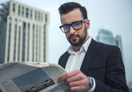 Handsome businessman in suit and glasses is reading newspaper while standing on the balconyの写真素材