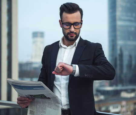 Handsome businessman in suit and glasses is reading newspaper and looking at his watch while standing on the balconyの写真素材