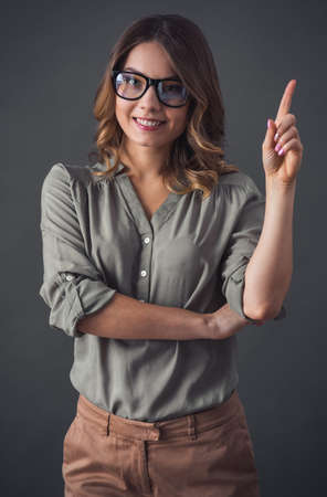 Attractive young woman in smart casual clothes and glasses is pointing upward, looking at camera and smiling, on gray backgroundの写真素材