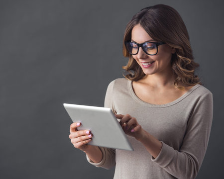 Attractive young woman in casual clothes and glasses is using a digital tablet and smiling, on gray...