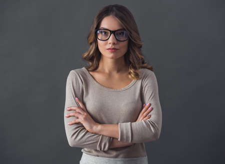 Attractive young woman in casual clothes and glasses is looking at camera while standing with crossed arms on gray backgroundの写真素材