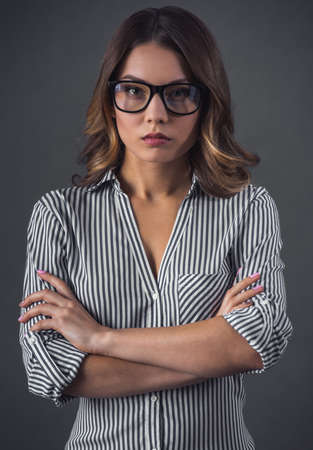 Attractive young woman in casual clothes and eyeglasses is looking at camera while standing with crossed arms, on gray backgroundの写真素材
