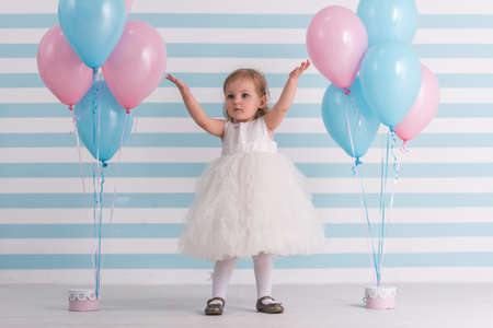 Cute little girl in fluffy white dress is raising hands up while standing near balloons, on light backgroundの写真素材