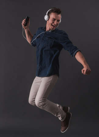 Handsome young man in headphones is listening to music and singing while jumping on gray backgroundの写真素材