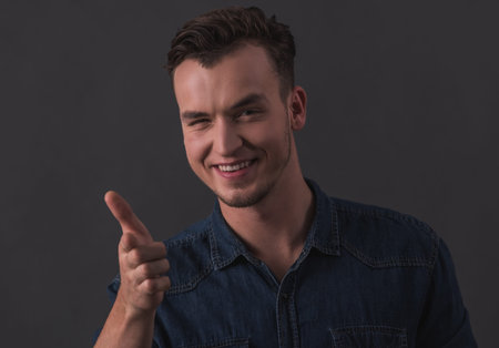Handsome young man is pointing, looking at camera and smiling, on gray background