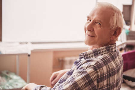 Handsome old man is looking at camera and smiling while sitting in wheelchair in hospital wardの写真素材