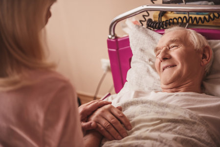 Cheerful old man is looking at his wife and smiling while lying in bed in the hospital ward
