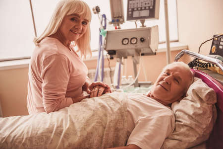 Cheerful old couple is looking at camera and smiling, man is lying in bed in the hospital wardの写真素材