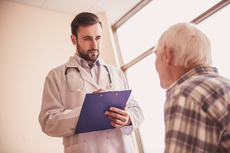 Handsome doctor is talking with an old man and making notes while standing in hospital wardの写真素材