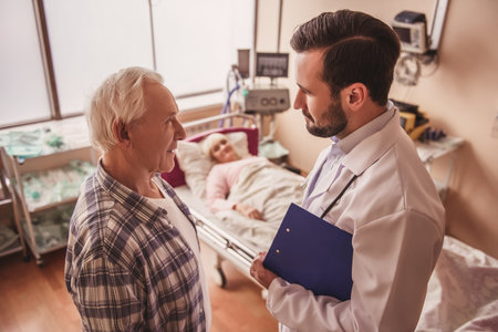 Old woman is sleeping in bed in the hospital ward while her husband and doctor are talking in the...