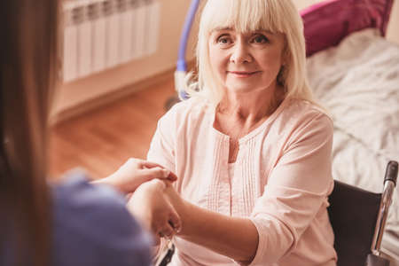 Beautiful old lady is holding doctor's hand and smiling while sitting in the wheelchair in hospital wardの写真素材