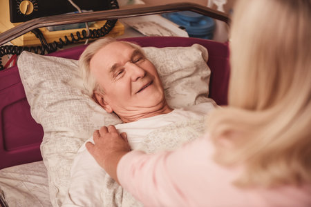 Cheerful old man is looking at his wife and smiling while lying in bed in the hospital ward