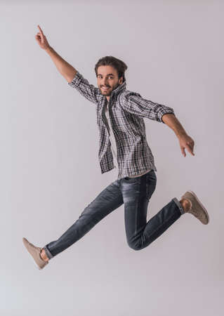Handsome bearded man in casual clothes is pointing away, jumping and smiling, on light backgroundの写真素材