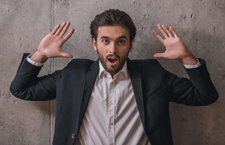 Handsome confident businessman with bristle, in suit, is holding hands up, mouth open and looking at camera, on gray backgroundの写真素材