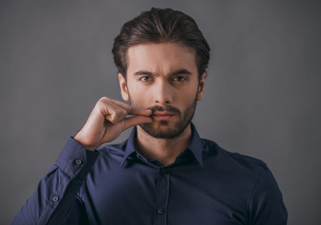 Handsome confident businessman in dark blue shirt is looking at camera and showing to keep the mouth closed, on gray backgroundの写真素材