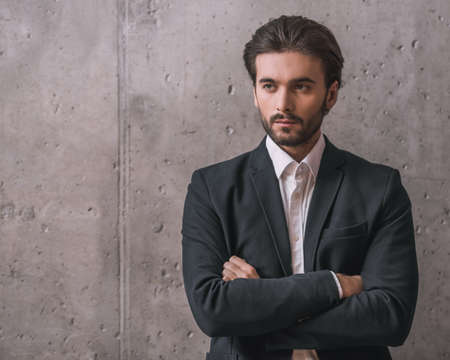 Handsome confident businessman with bristle, in suit, is standing with crossed arms on gray backgroundの写真素材