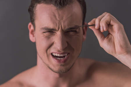 Handsome young man with bare torso is making a wry face while plucking eyebrows, on gray backgroundの写真素材
