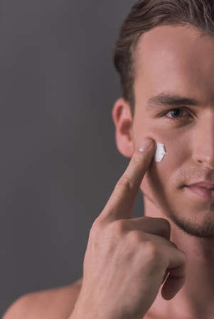 Cropped image of handsome young man applying face cream and looking at camera, on gray backgroundの写真素材