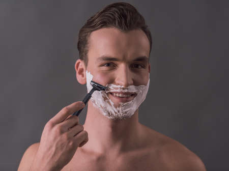 Handsome young man is shaving using a razor, looking at camera and smiling, on gray backgroundの写真素材