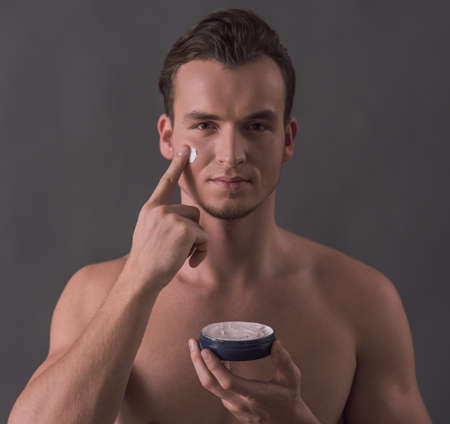Handsome young man is applying face cream and looking at camera, on gray backgroundの写真素材