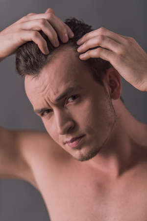 Handsome young man with bare torso is examining his hair and looking at camera, on gray backgroundの写真素材