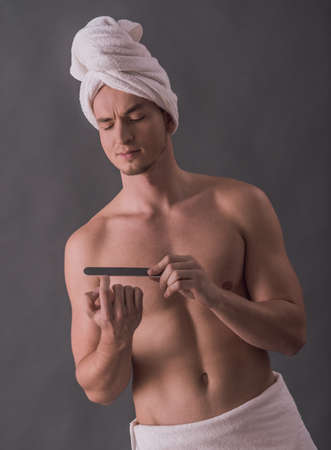 Handsome young man with bare torso, towel on his thighs and head is filing fingernails, on gray backgroundの写真素材