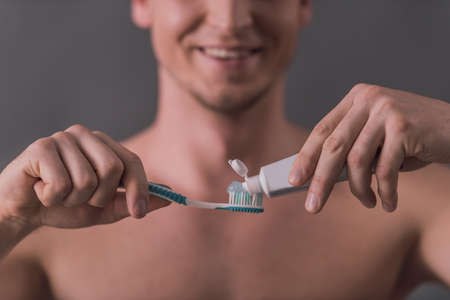 Cropped image of handsome young man applying toothpaste on a toothbrush and smiling, on gray backgroundの写真素材