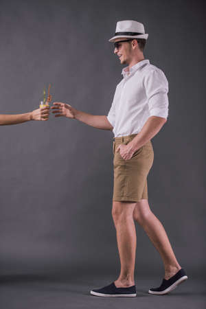 Full-length, back view image of handsome young man in shirt, shorts, hat and sun glasses taking a cocktail and smiling while walking on gray backgroundの写真素材