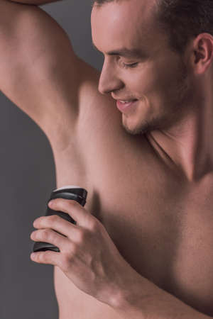Handsome young man with bare torso is using a stick antiperspirant and smiling, on gray backgroundの写真素材