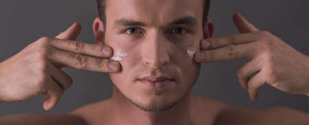 Handsome young man is applying face cream and looking at camera, on gray backgroundの写真素材