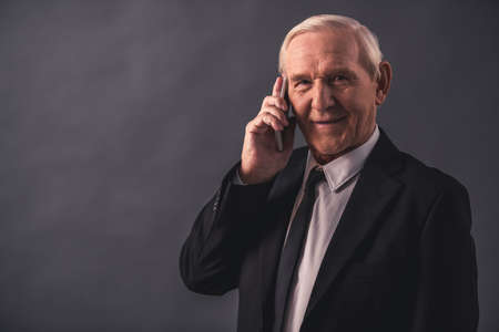 Handsome old businessman in suit is talking on the mobile phone, looking at camera and smiling, on gray backgroundの写真素材