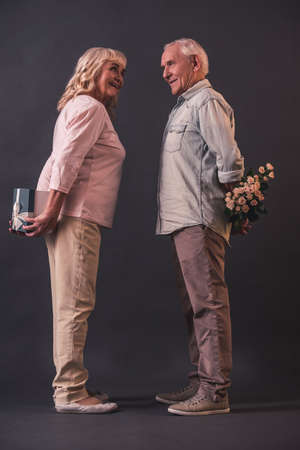 Beautiful old man and woman in casual clothes are holding presents for each other behind their backs and smiling, on gray backgroundの写真素材