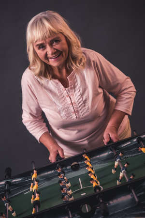 Beautiful old woman in casual clothes is playing table football, looking at camera and smiling, on gray backgroundの写真素材