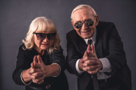 Beautiful old couple in elegant clothes and glasses is holding hands together imitating guns, looking at camera and smiling, on gray backgroundの写真素材