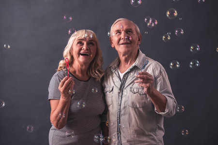 Beautiful old man and woman in casual clothes are blowing soap bubbles and smiling, on gray...