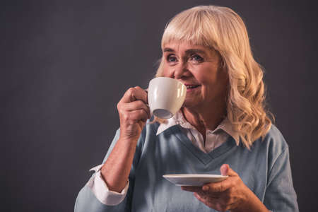 Beautiful old lady in casual clothes is holding a cup and smiling, on gray backgroundの写真素材