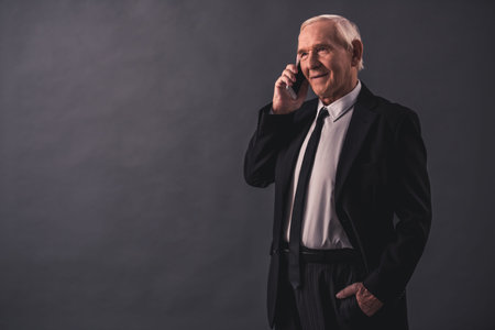 Handsome old businessman in suit is talking on the mobile phone and smiling, on gray background