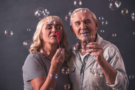 Beautiful old man and woman in casual clothes are blowing soap bubbles and smiling, on gray backgroundの写真素材