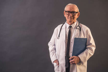 Handsome old doctor in medical coat and eyeglasses is holding a folder and looking at camera, on gray backgroundの写真素材