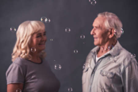 Beautiful old man and woman in casual clothes are looking at each other and smiling, on gray backgroundの写真素材