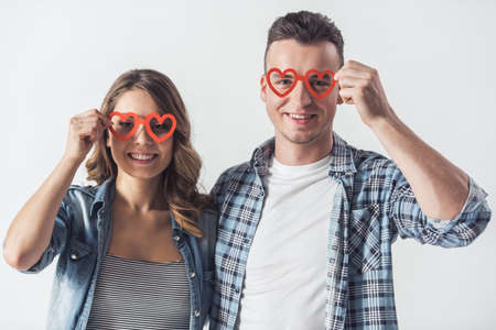 Beautiful young couple in casual clothes is holding two red heart-shaped paper glasses, looking at camera and smiling, isolated on whiteの写真素材