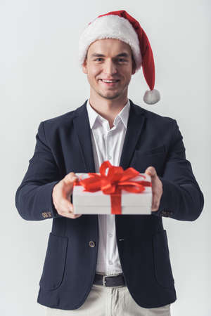 Handsome guy in suit and Santa hat is holding a gift box, looking at camera and smiling, isolated on whiteの写真素材