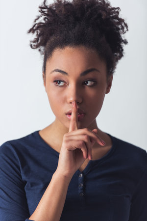 Beautiful Afro-American girl in casual clothes is looking away and showing silence sign, isolated on...