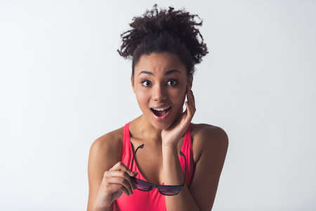 Beautiful Afro-American girl in bright pink swimsuit is holding sun glasses and looking at camera with the open mouth, isolated on whiteの写真素材