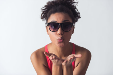 Beautiful Afro-American girl in bright pink swimsuit and sun glasses is sending air kiss at camera,...