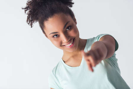 Cheerful Afro-American girl in casual clothes is pointing and looking at camera and smiling, isolated on whiteの写真素材