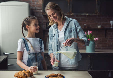 Mom and daughter in casual clothes are  hugging and smiling while mom is pouring milk, in kitchen at homeの写真素材