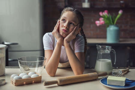 Cute little girl in apron is leaning on the kitchen table and thinking what to bakeの写真素材
