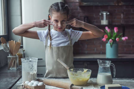 Cute little girl in apron is making lines on her face with flour ready to cook, in kitchen at homeの写真素材