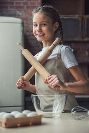 Cute little girl in apron is holding two crossed rolling pins, looking at camera and smiling while cooking in kitchen at homeの写真素材